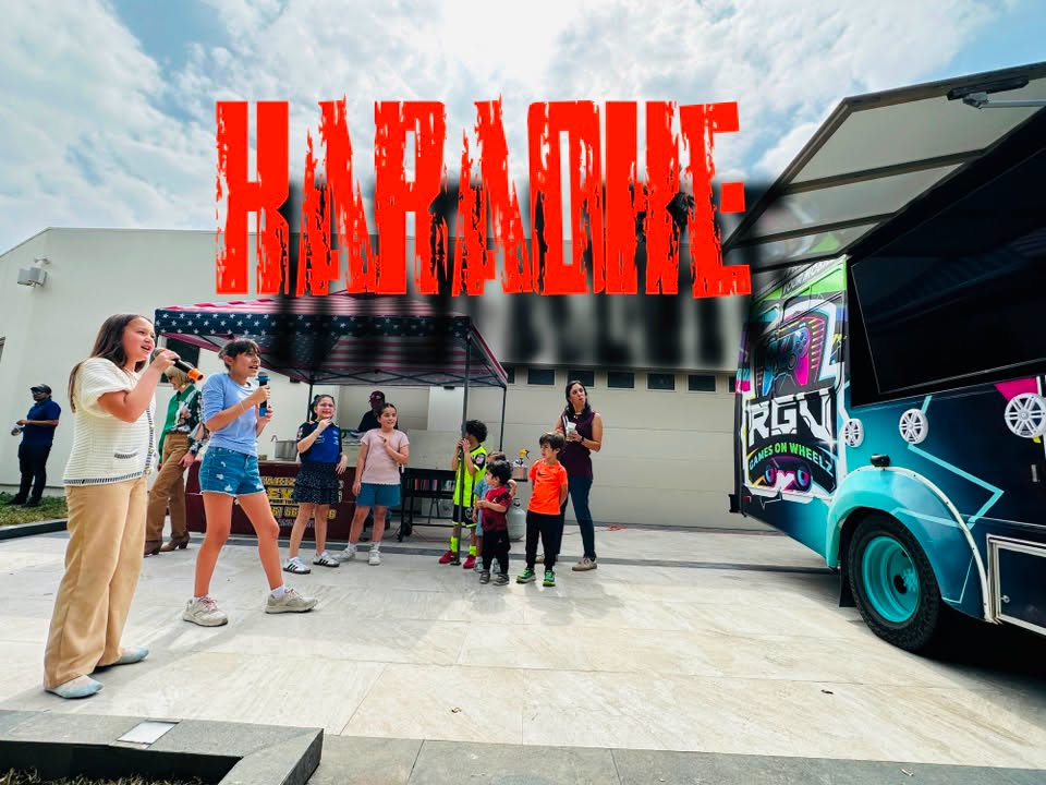 Two guests singing into microphones at an outdoor RGV Games on Wheelz karaoke setup, with the gaming bus parked alongside and guests watching